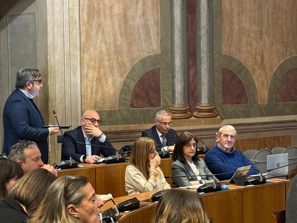 A group of people seated at a conference table in a meeting room, with one person standing and speaking. The setting features embellished walls and a formal atmosphere.