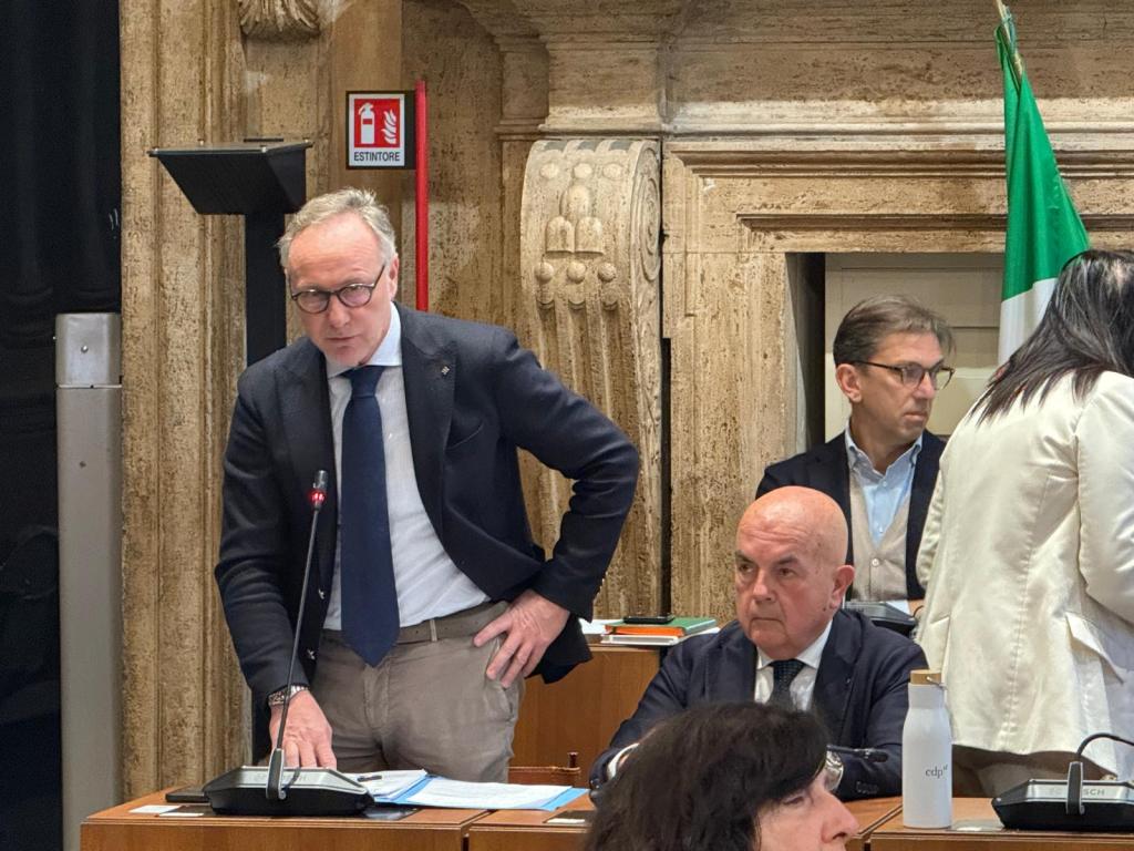 A man speaking at a podium in a formal setting, wearing glasses and a suit, with a microphone in front of him. Two other men seated nearby are listening attentively, and a woman stands behind them. The background features a stone wall and an Italian flag.