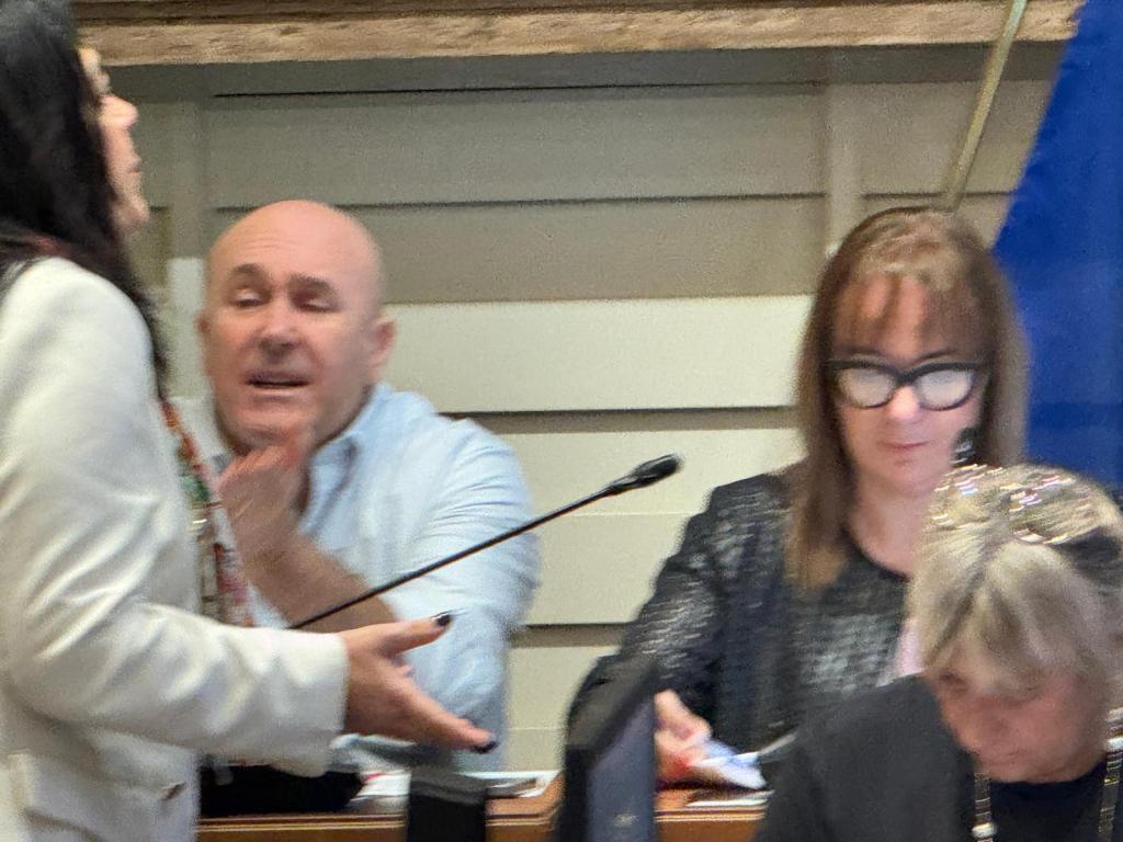 A meeting scene featuring a woman in a white jacket speaking animatedly, a man in a blue shirt gesturing in response, and a woman with glasses focused on paperwork in the background.
