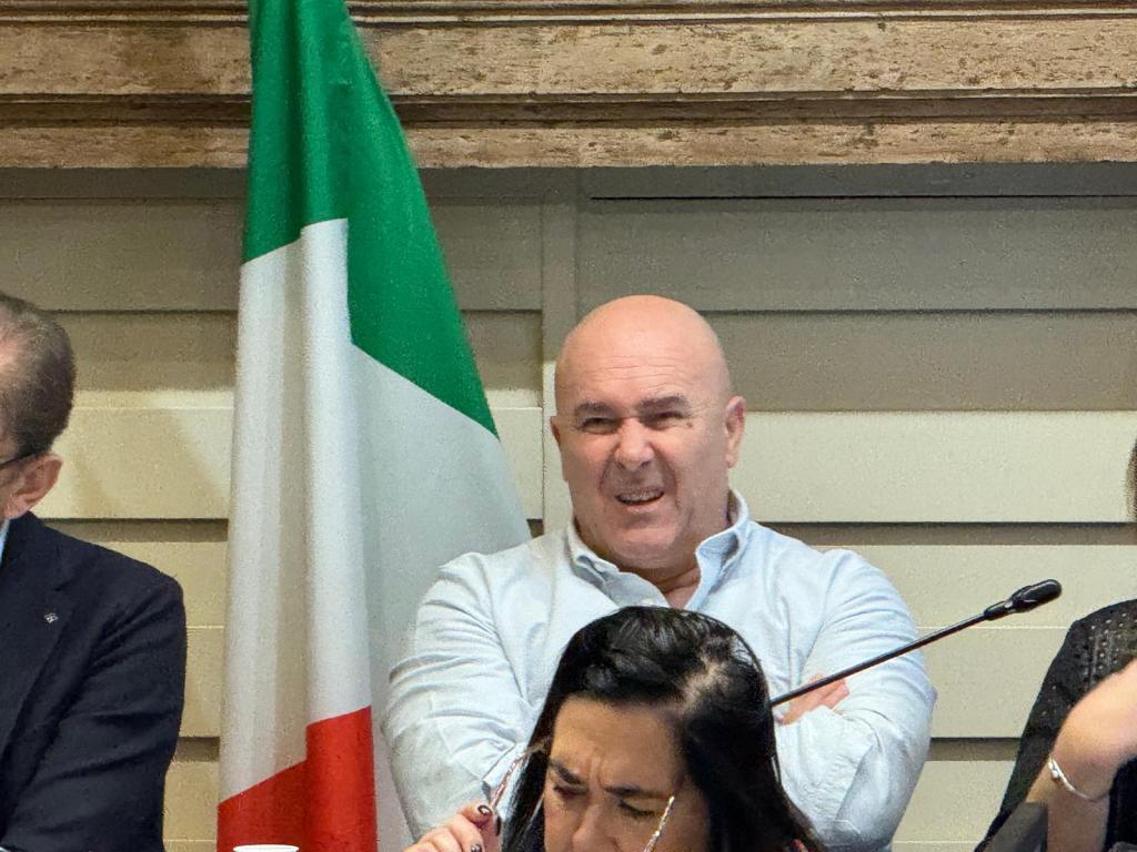 Man making a facial expression of discontent or confusion, seated in a meeting with a blurred woman in front of him and an Italian flag in the background.