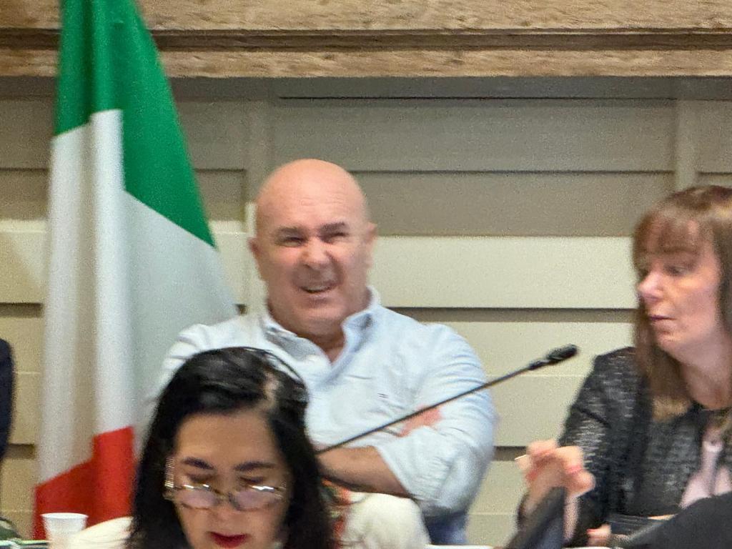 A group of people in a meeting setting, with a visible Italian flag in the background. A man is frowning while sitting next to a woman who is speaking.