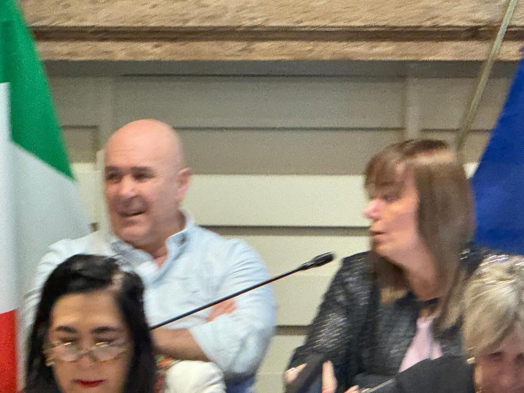 Two people engaged in discussion at a conference table, with the Italian flag in the background.
