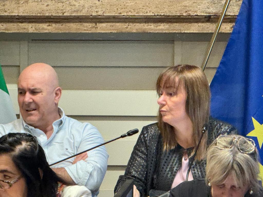A woman and a man seated at a table during a meeting, with the woman looking thoughtfully and the man expressing concern. In the background, there are flags, including an Italian flag and an EU flag.