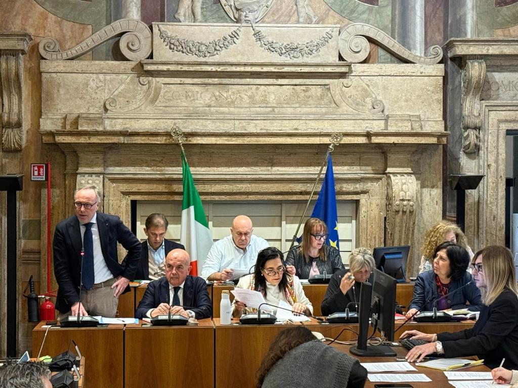 A group of officials engaged in a meeting at a formal setting, with an Italian flag and an EU flag displayed. The table is cluttered with papers and electronic devices as they discuss matters.