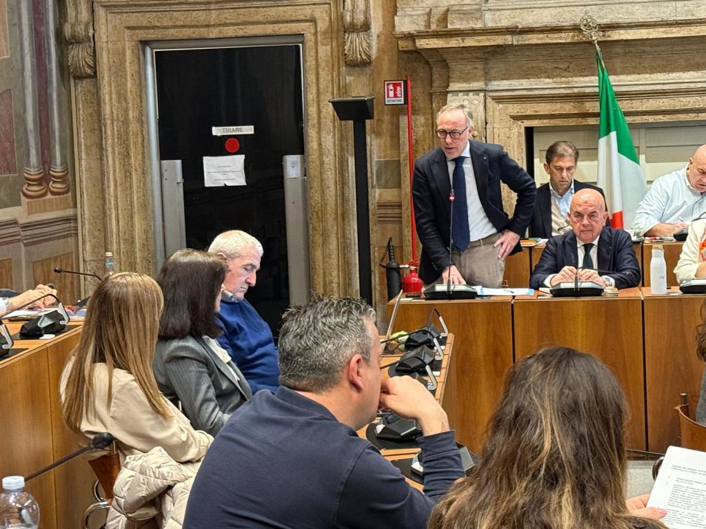 A meeting taking place in a formal setting with several individuals seated at a table, while one man stands and speaks. The room features ornate walls and a visible Italian flag.