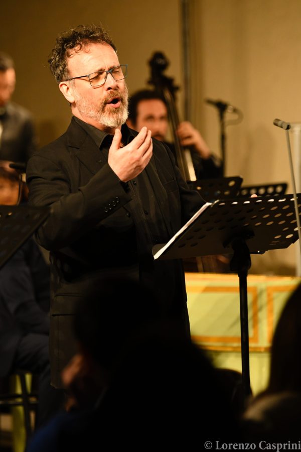 A man with curly hair and glasses passionately singing on stage, dressed in black. He gestures with his hand while performing, with musicians visible in the background.