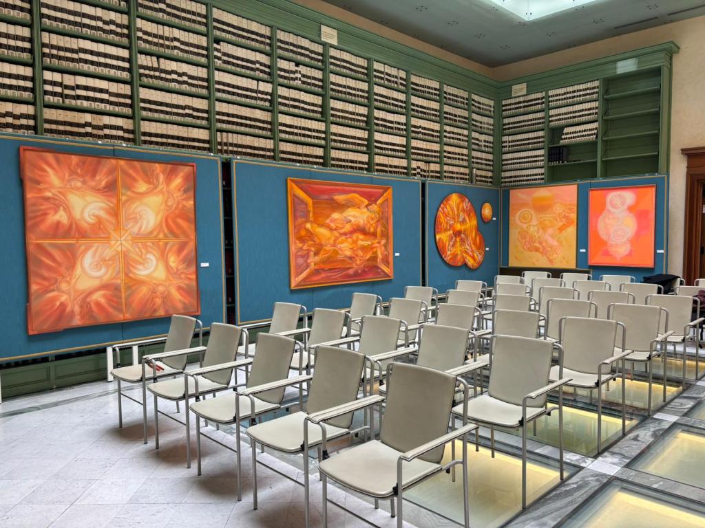 An art gallery featuring vibrant, abstract paintings in shades of orange, displayed on blue boards against a backdrop of bookshelves. Rows of modern, silver-framed chairs are arranged on a marble floor, ready for visitors.