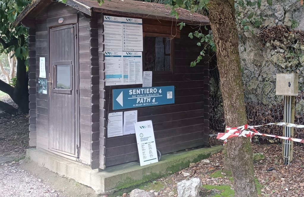 A small wooden hut with a sign indicating 'Sentiero 4' and 'Path 4,' surrounded by trees and red and white caution tape.