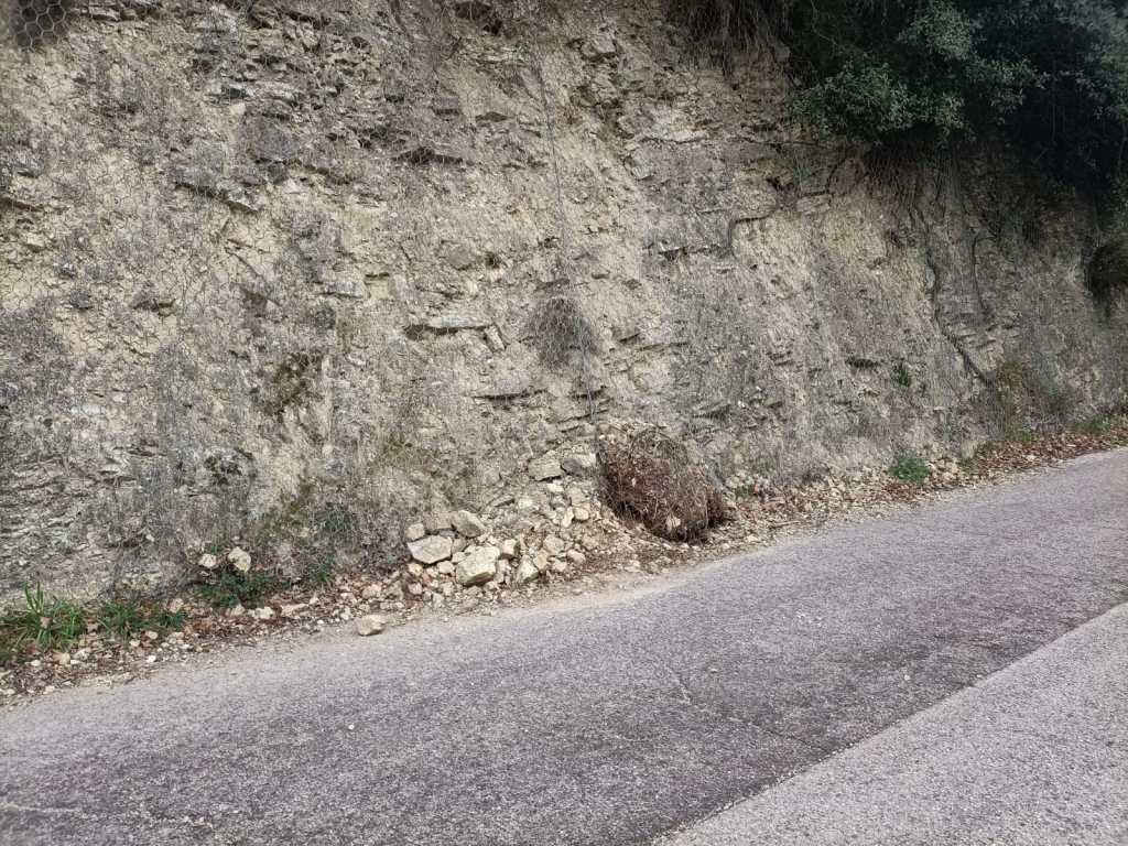 A roadside view featuring a rocky cliffside with scattered stones and patches of vegetation next to a paved road.