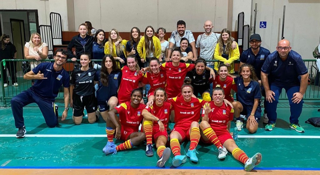 A group photo of a women's football (soccer) team celebrating, wearing red jerseys with a team logo, posing with coaches and supporters in a gym or sports hall.