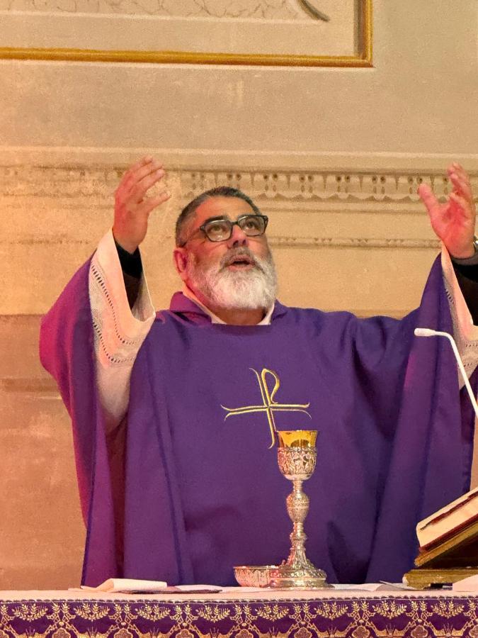 A priest in a purple vestment raises his arms in a gesture of blessing during a religious service, with a chalice and a Bible on the altar in front of him.