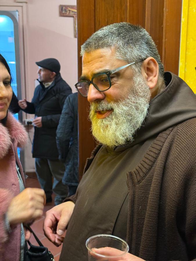 A man with a grey beard and glasses wearing a brown robe is talking to a woman with dark hair and a pink fur coat. They are in an indoor setting, and there are other people in the background.