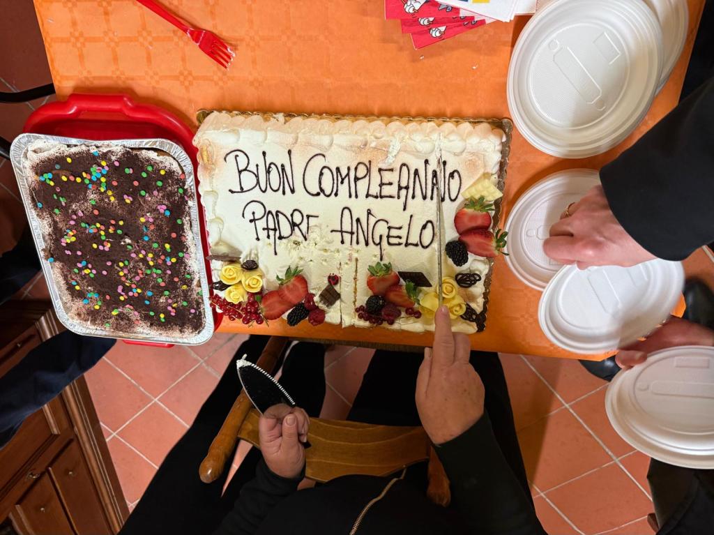 A birthday cake decorated with fruits and the inscription 'BUON COMPLEANNO PADRE ANGELO' on a table, alongside a tray of dessert with colorful sprinkles.