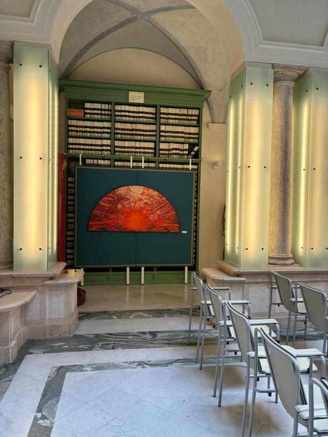 A large artwork featuring a red sun and abstract patterns is displayed on a green backdrop within a spacious architectural setting. To the left, a tall bookshelf filled with boxes can be seen, and several metal chairs are arranged in front.