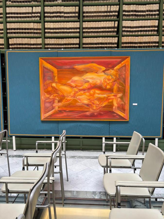 An abstract painting featuring warm tones of orange and red, displayed on a blue wall in a library setting, with rows of bookshelves filled with books in the background. Several chairs are arranged in front of the artwork.