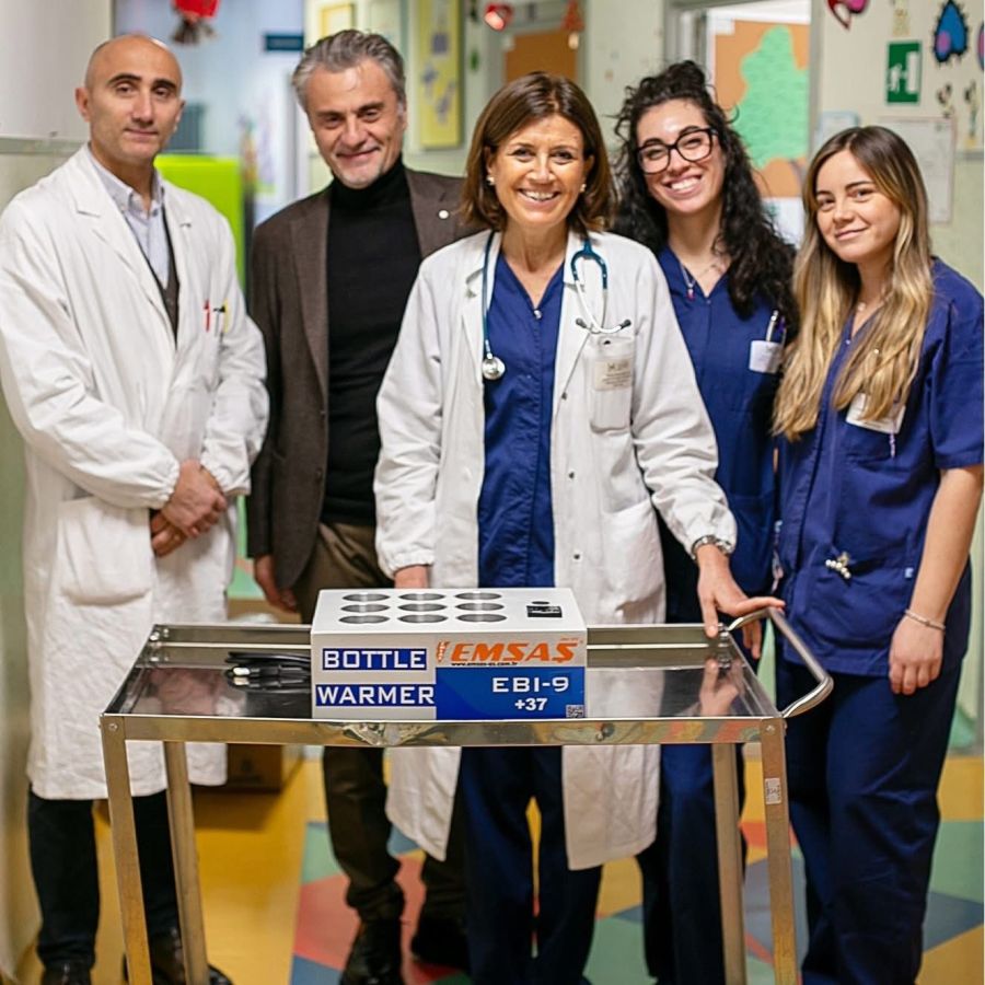 A group of healthcare professionals, including four individuals wearing scrubs and one in a white coat, poses in a hospital setting beside a bottle warmer on a metal cart.