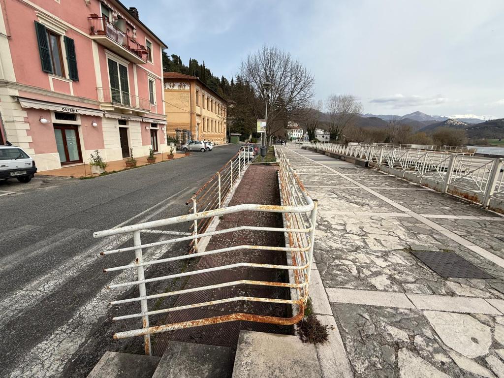 View of a street with a pink building on the left, a rusty railing along a pathway, and mountains in the background under a cloudy sky.