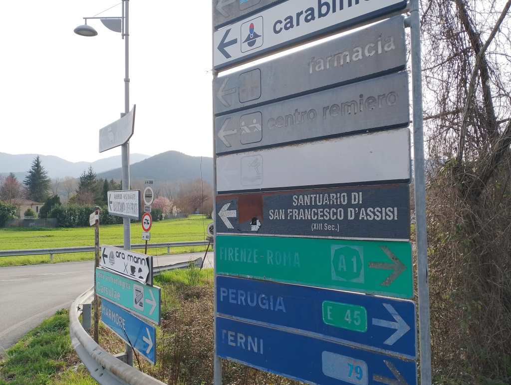 A collection of road signs in a rural area indicating directions to various destinations, including 'FIRENZE-ROMA', 'PERUGIA', and 'TERNI', along with local services and attractions.
