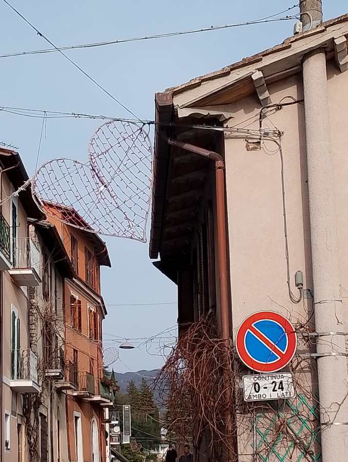 A scenic street view featuring two buildings with colorful facades. A heart-shaped decoration is suspended between them, and a no-parking sign is visible on the right side.