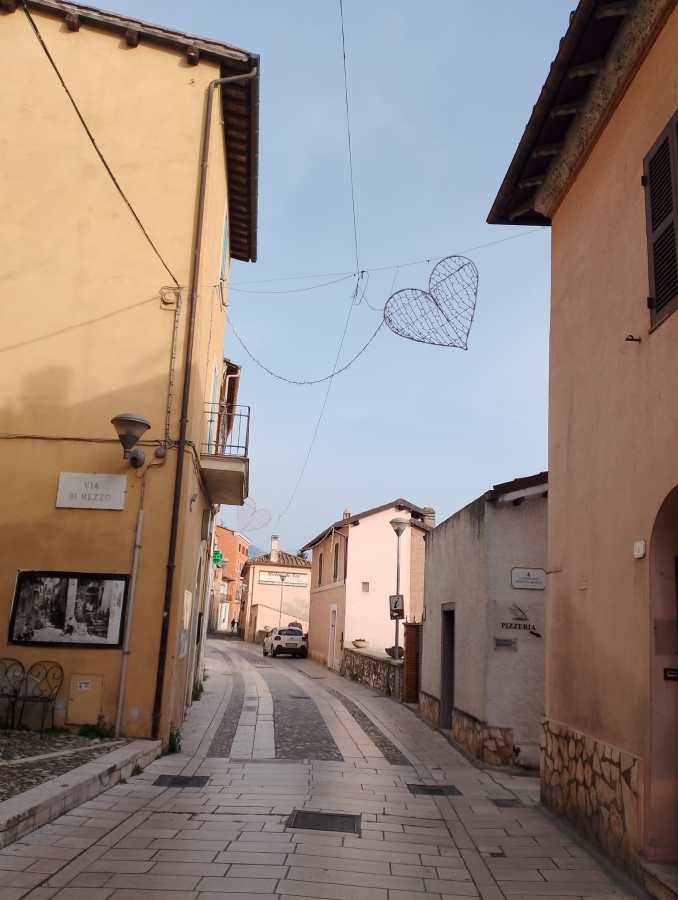 A narrow street in a quaint village featuring pastel-colored buildings, a decorative heart hanging overhead, and a parked car.