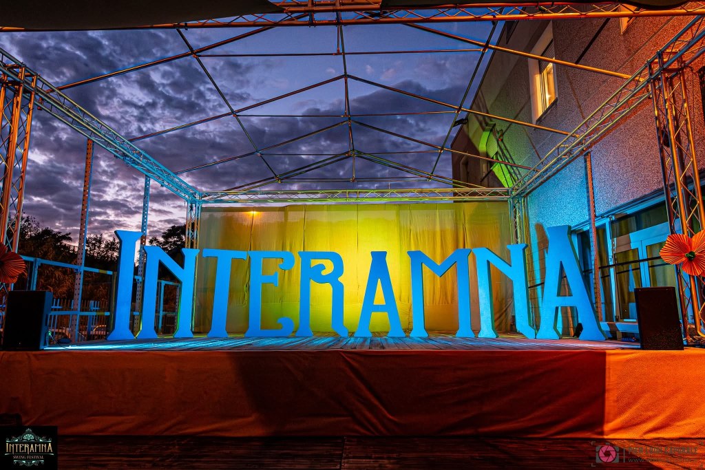 A stage setup for an event featuring large blue letters spelling 'INTERAMNA', illuminated by colorful lights against a twilight sky.