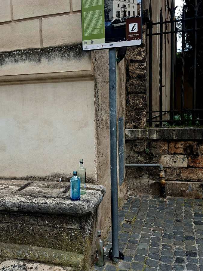 A weathered wall with a green and brown information sign attached to a metal pole, beside a stone bench. There are three glass bottles, one blue, one clear, and one green, placed on the bench, with cobblestones in the foreground.