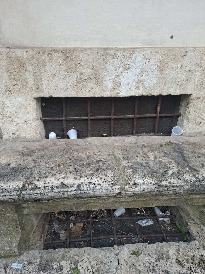A close-up view of a stone wall with a barred window at its base, containing discarded plastic cups and debris.