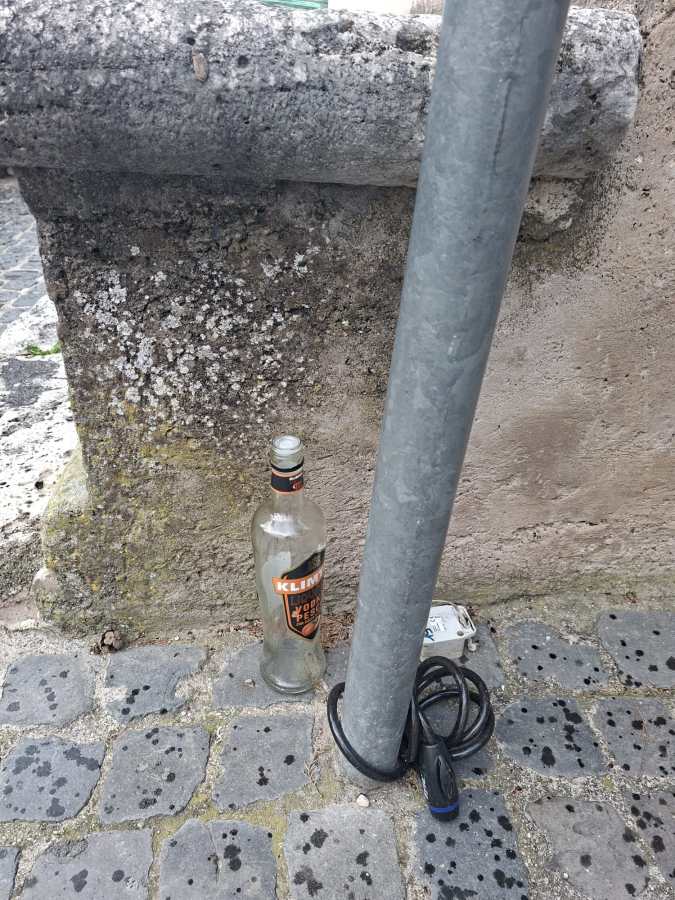 An empty glass bottle leaning against a concrete wall next to a metal pole, with a black cable tied to the pole, on a cobblestone street.