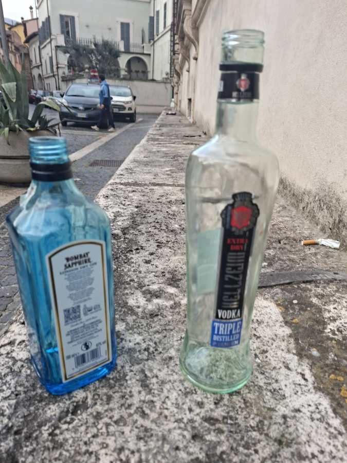 Two empty liquor bottles, one blue and one clear, sit on a stone surface in an outdoor setting, with a person walking by in the background.