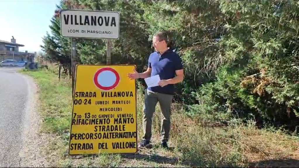 A man stands beside a yellow road sign indicating road maintenance on Strada Villanova, with a road closure notice and alternative route instructions. The background features green trees and a rural setting.