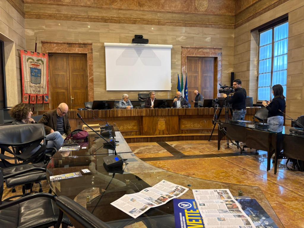 A meeting in a formal conference room with wooden decor, featuring several people seated at a table. A large screen is displayed in the background, along with flags. A camera is set up to capture the event.