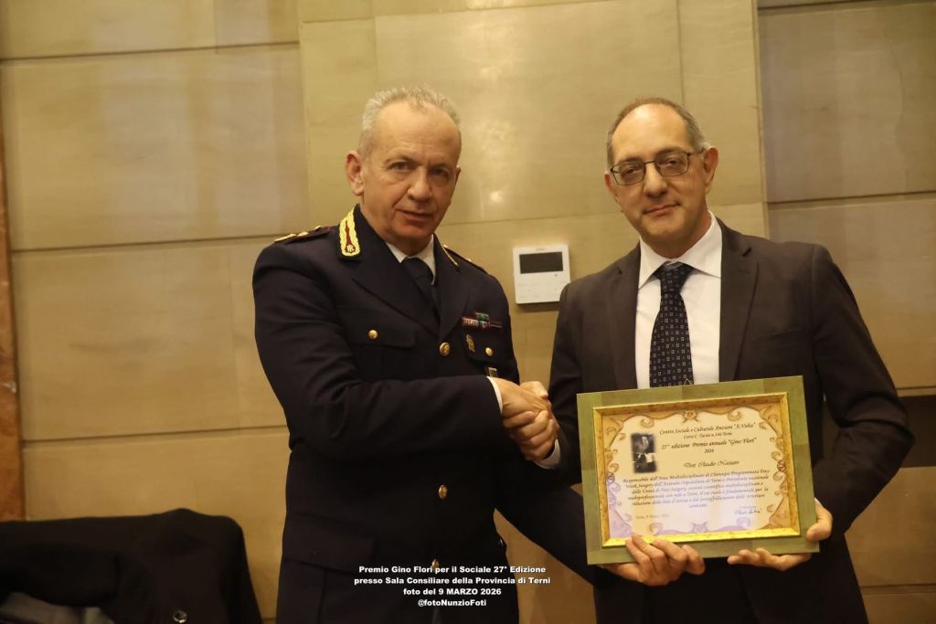 Two men shaking hands in a formal setting, one in a police uniform and the other in a suit, with a framed certificate in the latter's hand.