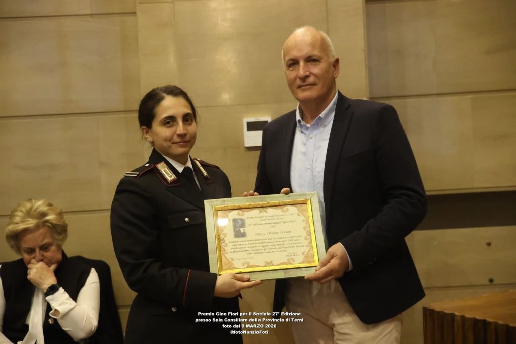 A female officer in uniform holds an award certificate alongside a man in formal attire, both appearing at a ceremony. A woman sits in the background, looking contemplative.