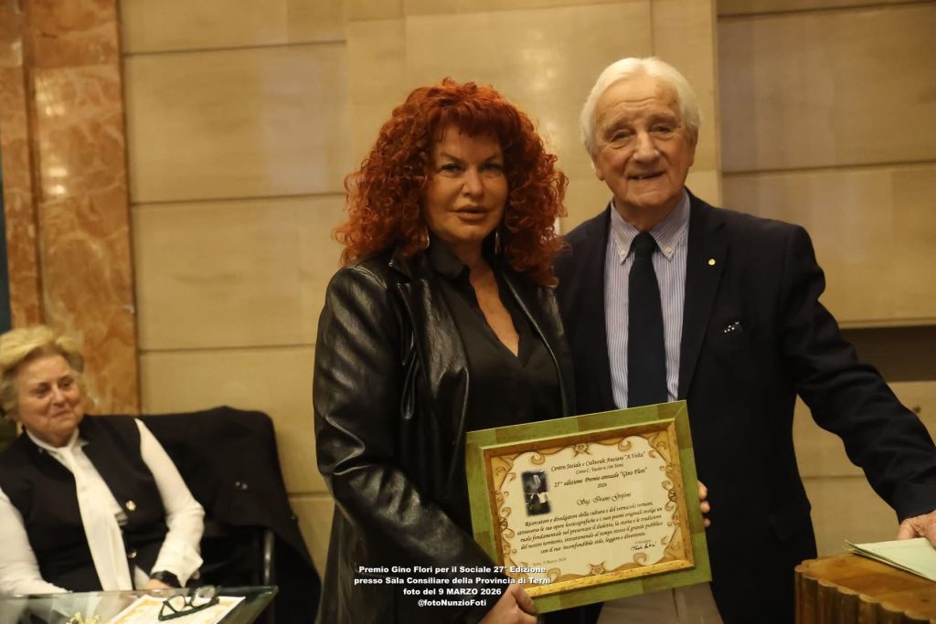 A woman with curly red hair and a black jacket stands next to an older man in a suit, both smiling while holding a framed award certificate. A woman can be seen sitting in the background.