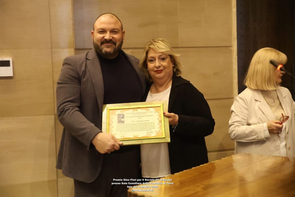 Two individuals are standing together, one holding a certificate. They appear to be at a formal event, possibly an award ceremony.
