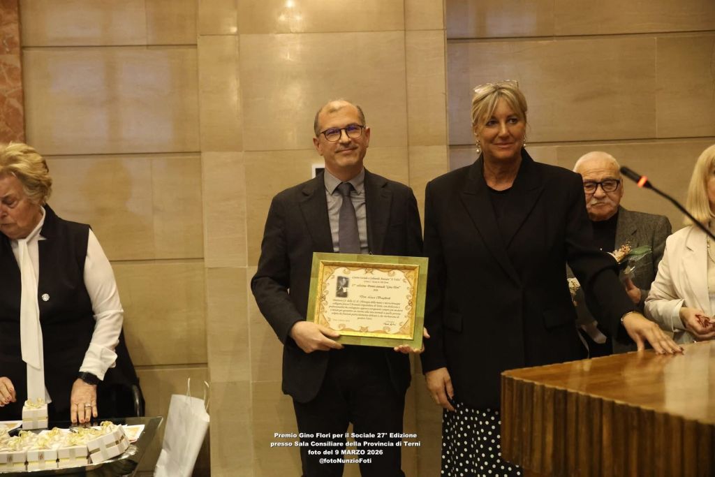A man in a suit holding a framed award stands next to a woman in a black outfit at a formal event. Other attendees are in the background, with gift boxes on a table.