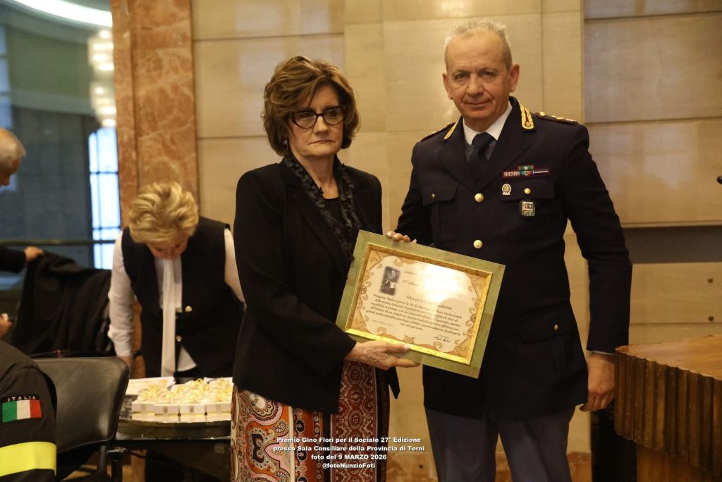 A formal event scene featuring a woman receiving a certificate from a man in uniform, with a table of snacks in the background. The setting appears to be an elegant hall.