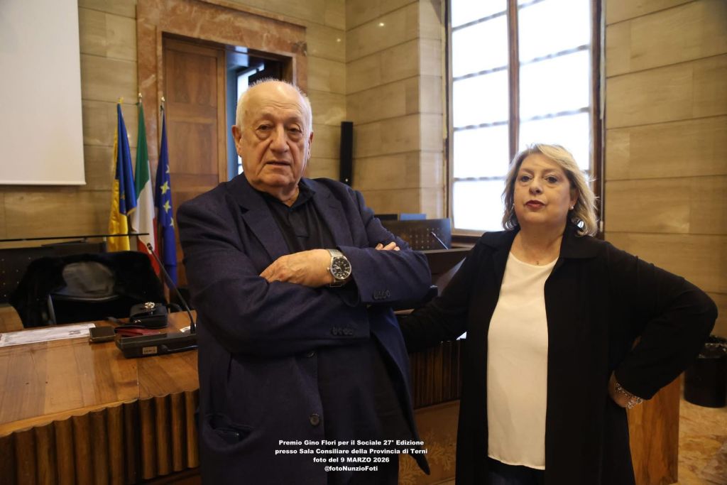A man and a woman pose together in a formal setting with flags in the background. The man has white hair and is wearing a dark blazer, while the woman has shoulder-length hair and is dressed in black.
