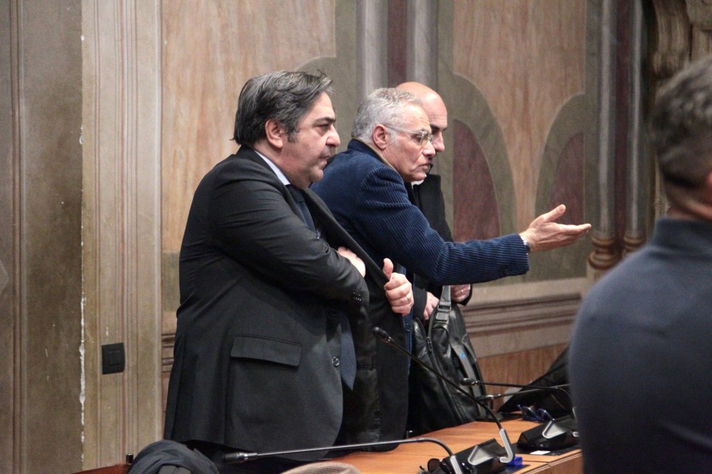 Three men standing at a conference table in a formal setting, engaged in conversation.