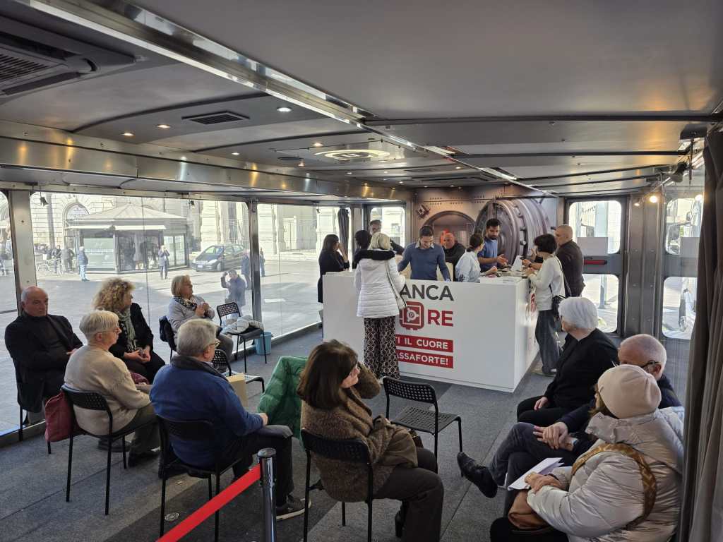 Inside a mobile bank setup, several people are seated in a waiting area while others interact at a service counter. The space features large windows and a modern design.