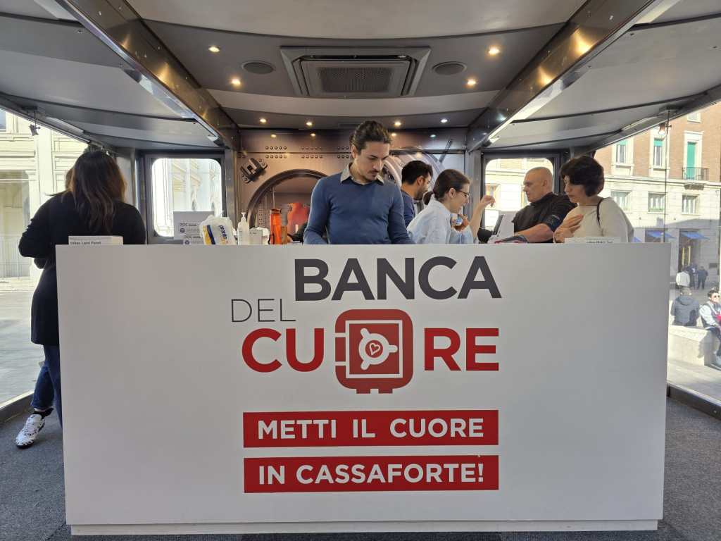 A modern banking setup with a white counter displaying the logo of 'Banca del Cuore.' People are gathered around the counter engaging in conversation. The background shows a view of the city.