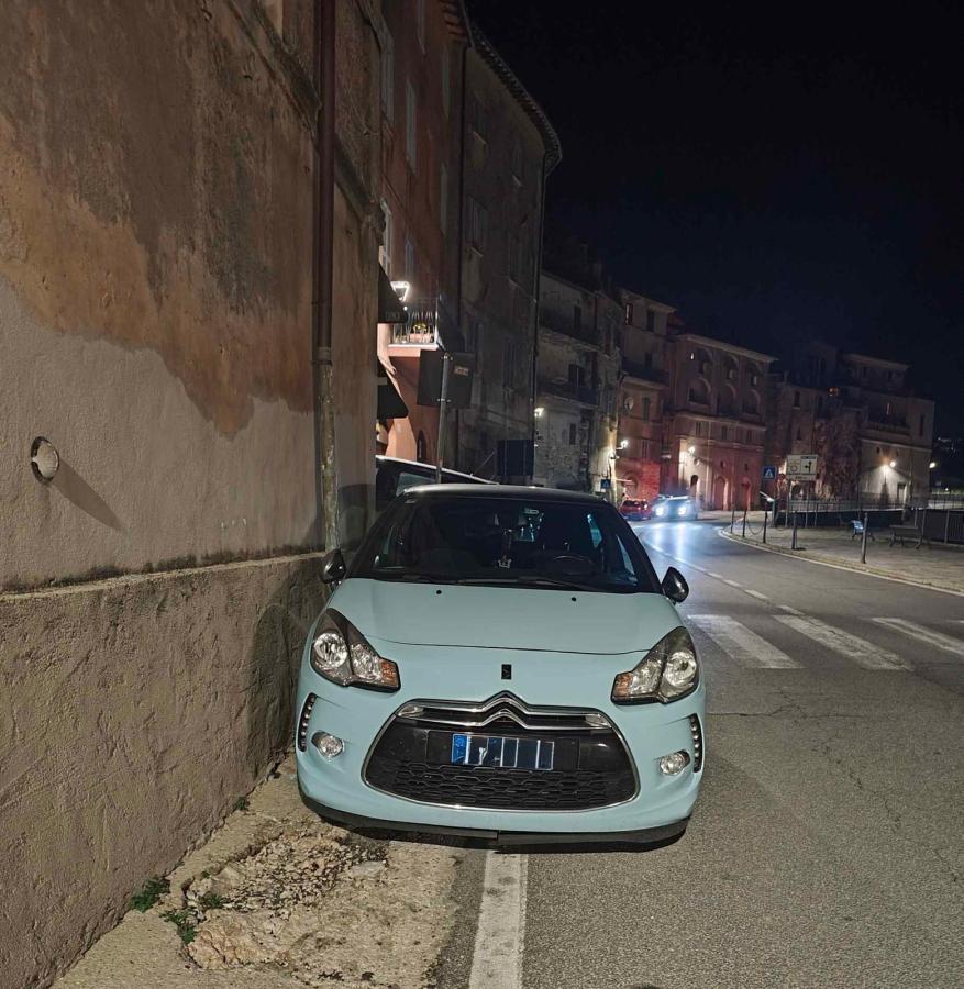 A light blue Citroën car parked closely against a wall in a dimly lit street at night, with buildings visible in the background.