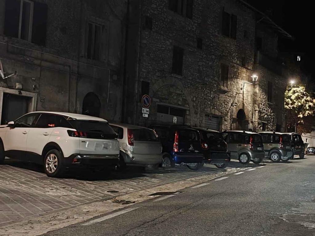 A row of parked cars on a cobblestone street at night, with stone buildings in the background and street lighting illuminating the scene.