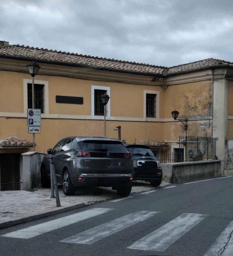 View of a street with two parked cars, one on a slope near a building with a yellow exterior and windows. Street signs visible indicate parking regulations. A cloudy sky is in the background.