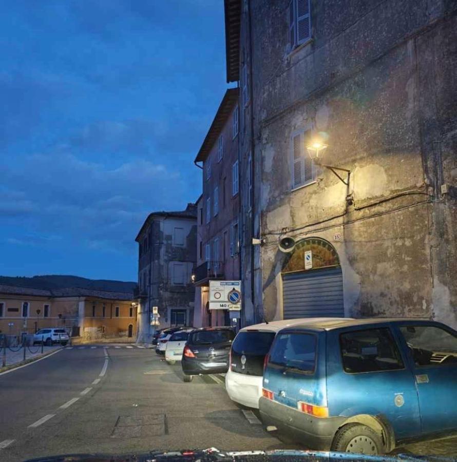 Street view at dusk featuring parked cars alongside a building with a weathered exterior and street lighting.