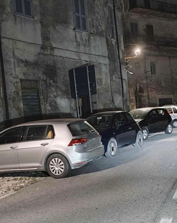 A row of parked cars on a narrow street at night, with a partially illuminated building in the background.