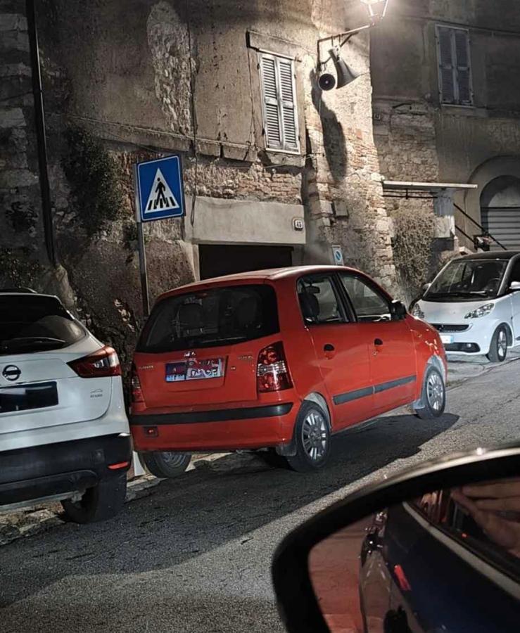 A red compact car parked between two other cars on a narrow street at night, with a pedestrian crossing sign visible in the background.