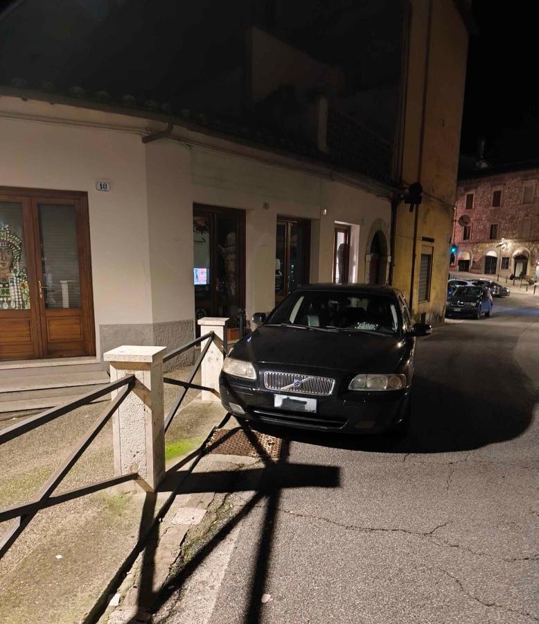 A parked black car on a narrow street at night, with a building and railing nearby.