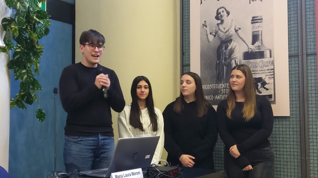 A young man speaking at a presentation with three women standing beside him, in front of a wall featuring an old advertisement.