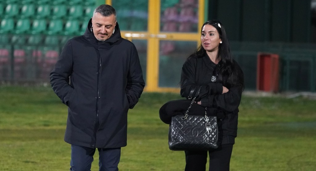 A man and a woman walking together on a football field. The man is wearing a dark coat and has his hands in his pockets, while the woman is dressed in black and holding a handbag. They appear to be engaged in conversation.